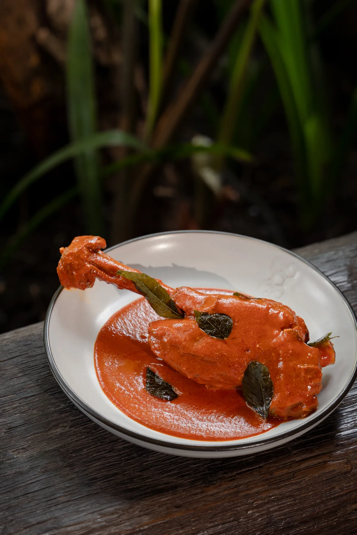 A braised dish plated with salam leaves in a pool of red sauce, on a wooden table in the garden