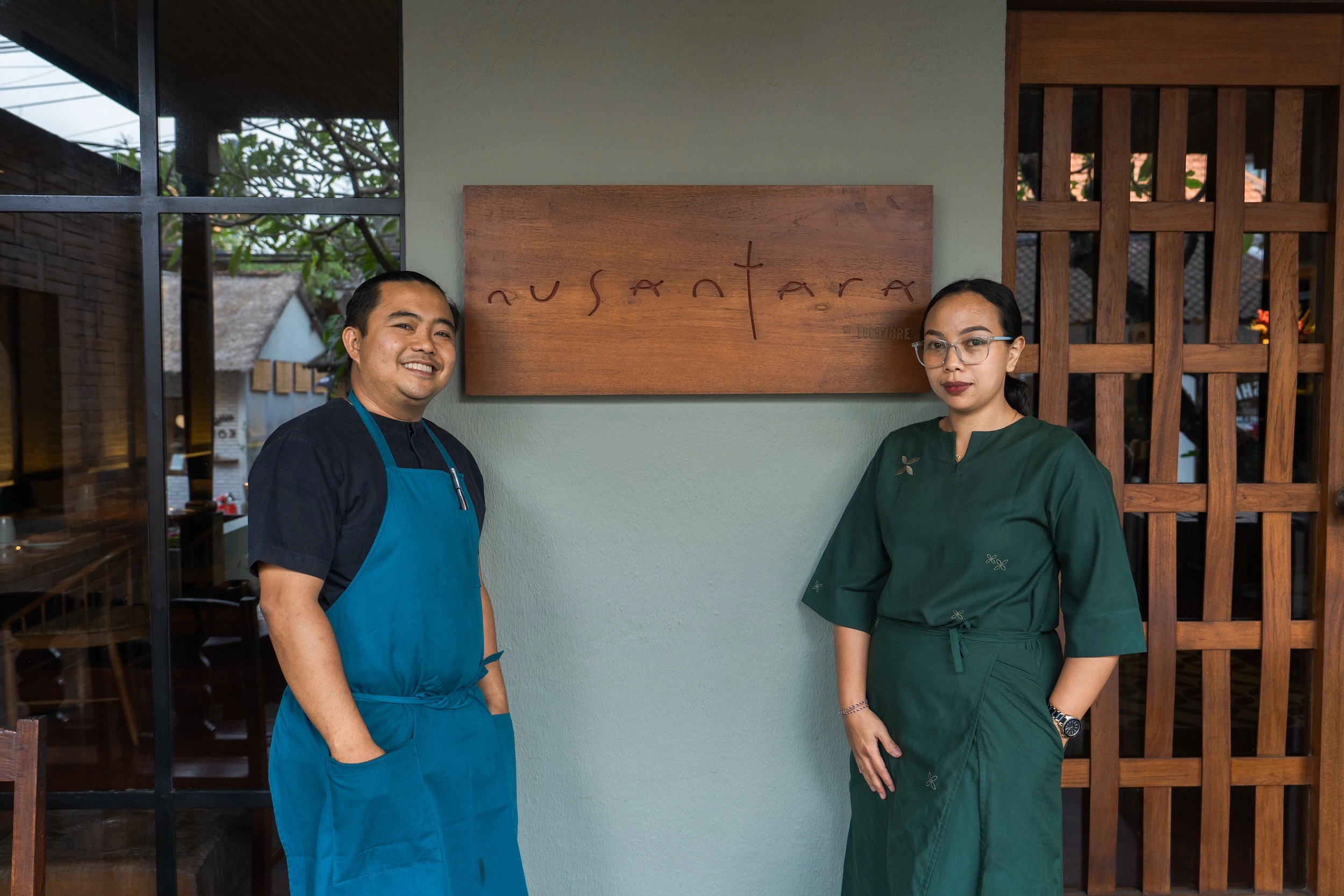 Chef Putu in a blue apron and Bu Wayan in a green dress, standing either side of the carved Nusantara nameplate