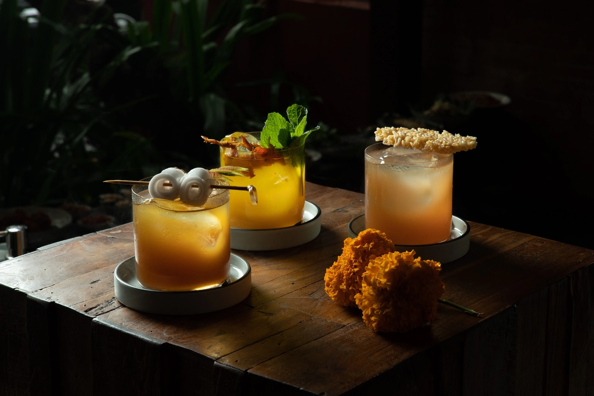 Three amber cocktails on a dark wooden table, garnished with mint, a skewer and a crisp wafer, beside two marigolds