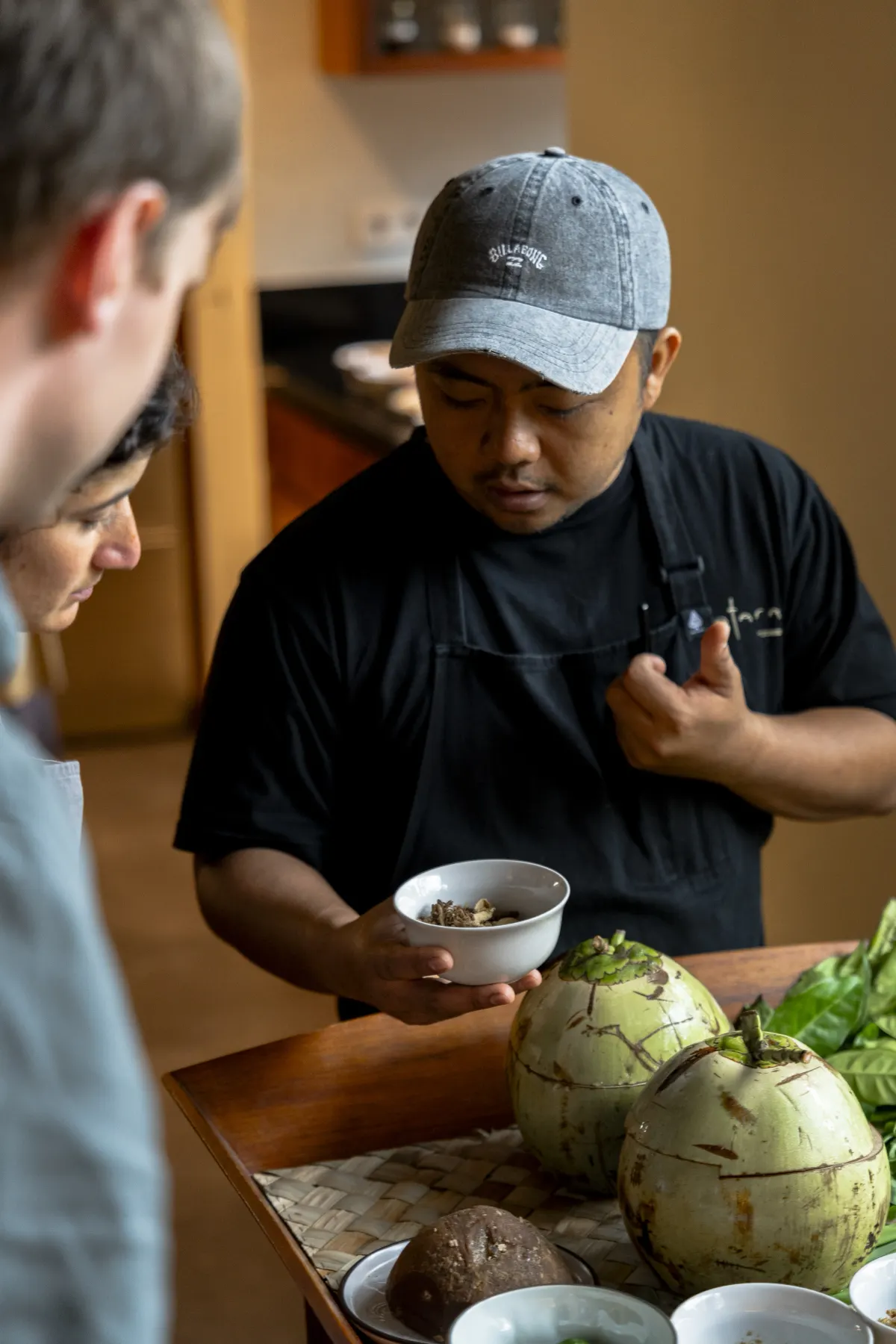 A chef talking a guest through an ingredient across a tray of young coconuts and herbs