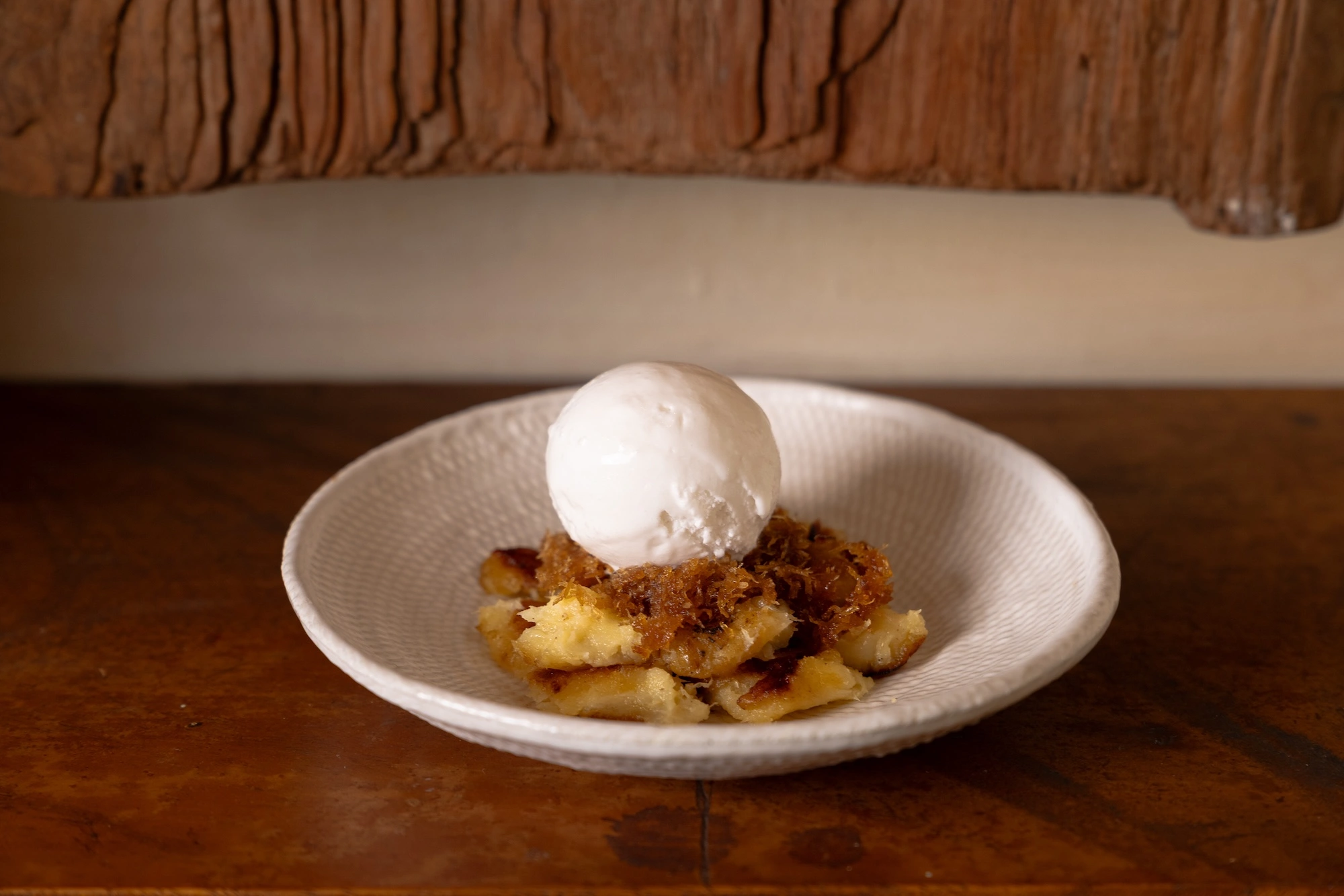 A scoop of white ice cream on caramelised fruit and a brown sugar crumb, in a textured ceramic bowl