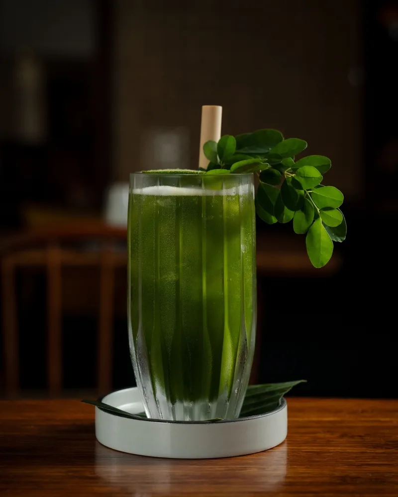 A tall glass of bright green kelor iced tea on a stone coaster, a sprig of moringa leaves over the rim