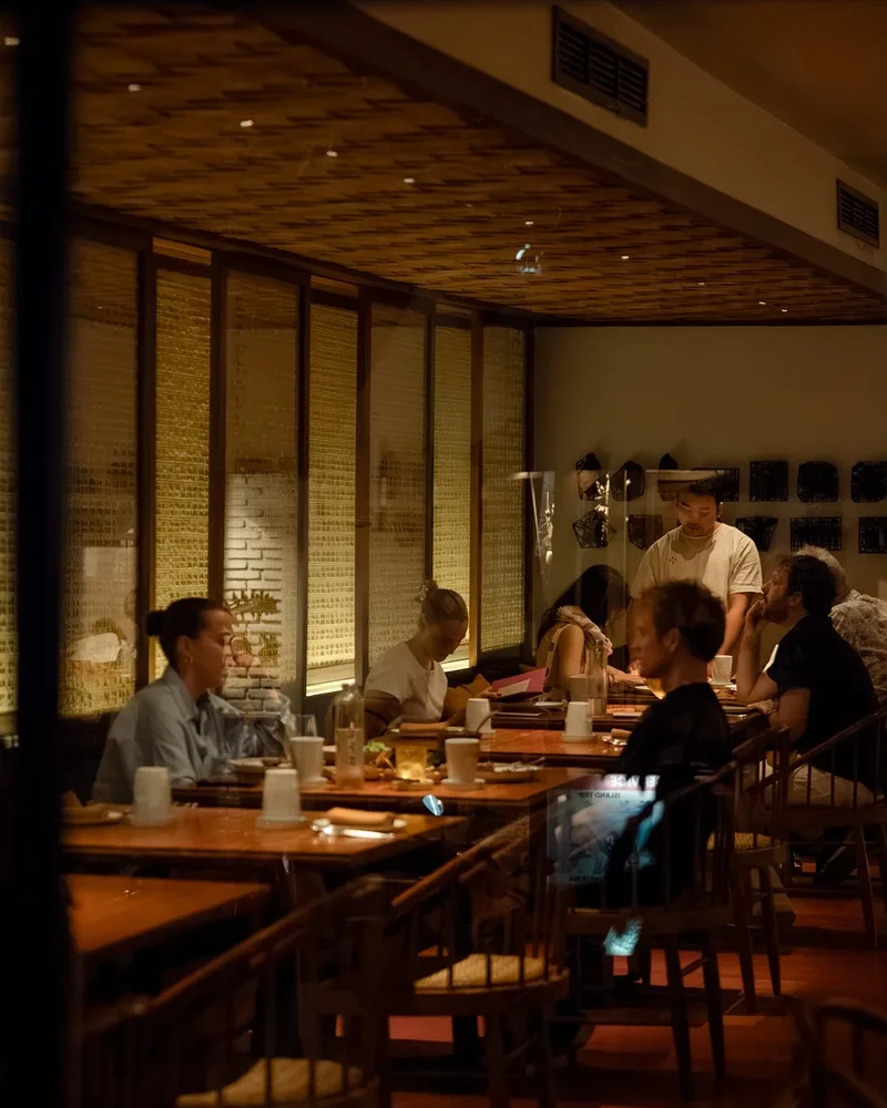 The dining room mid-service, guests along the long tables under warm light and rattan screens