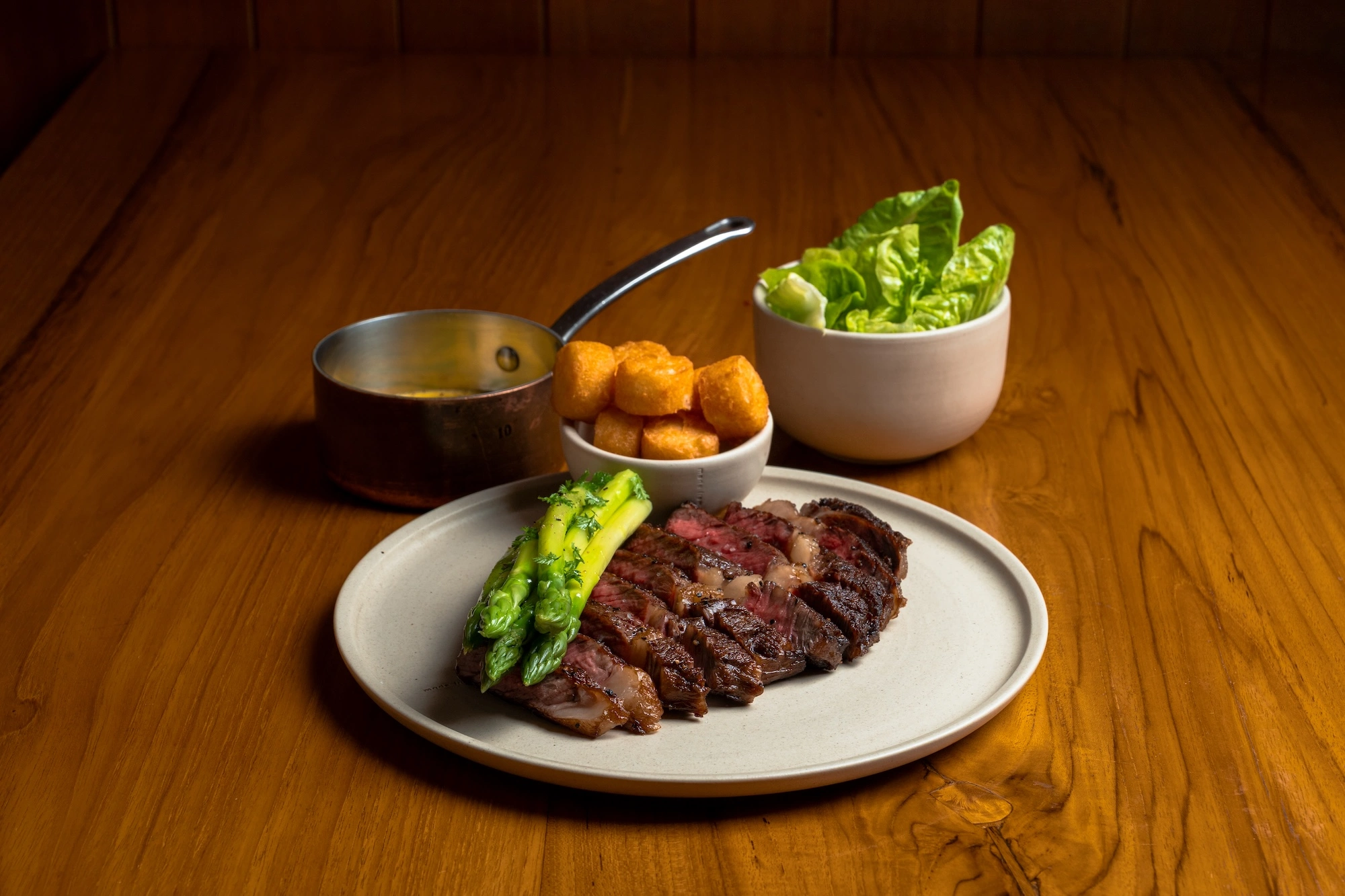 A sliced steak with grilled greens on a white plate, with a pan of sauce, a bowl of potato croquettes and a bowl of little gem lettuce behind, on a wooden table