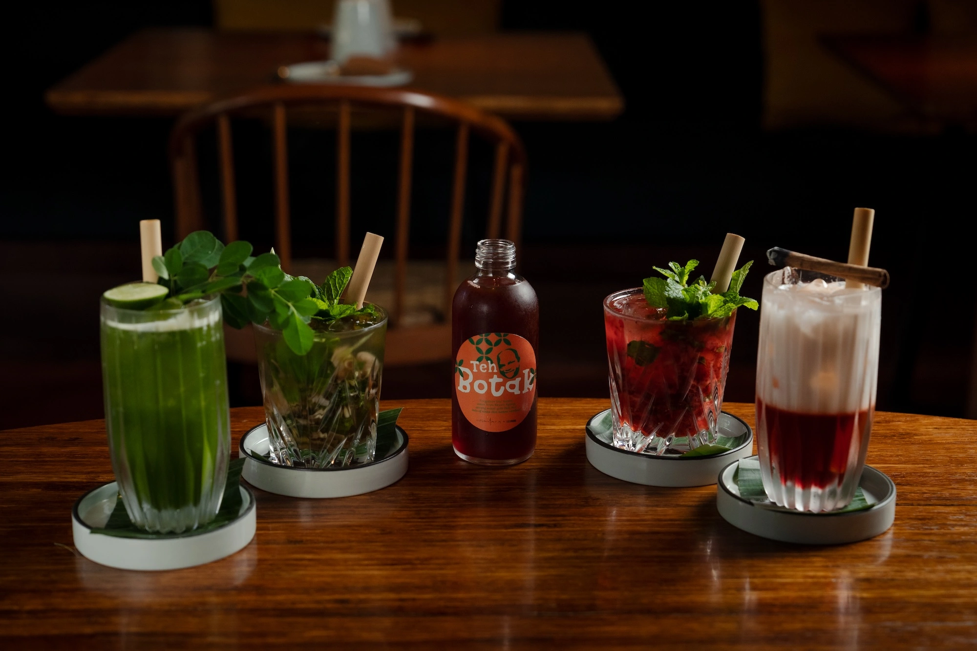 Five drinks on a wooden table — green and red ones topped with mint, a bottled drink, and a red one layered under white