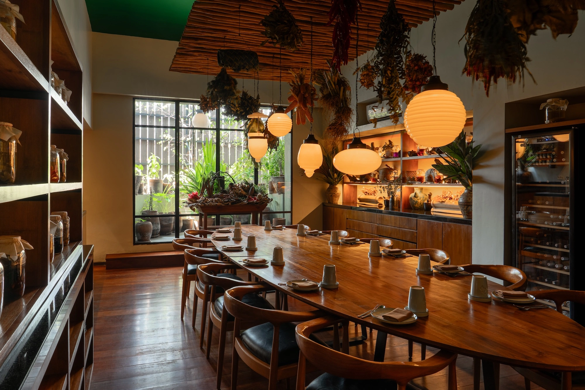 The private dining room — a long wooden table laid for a party under paper lanterns and hanging dried plants, with shelves of ferment jars along the wall