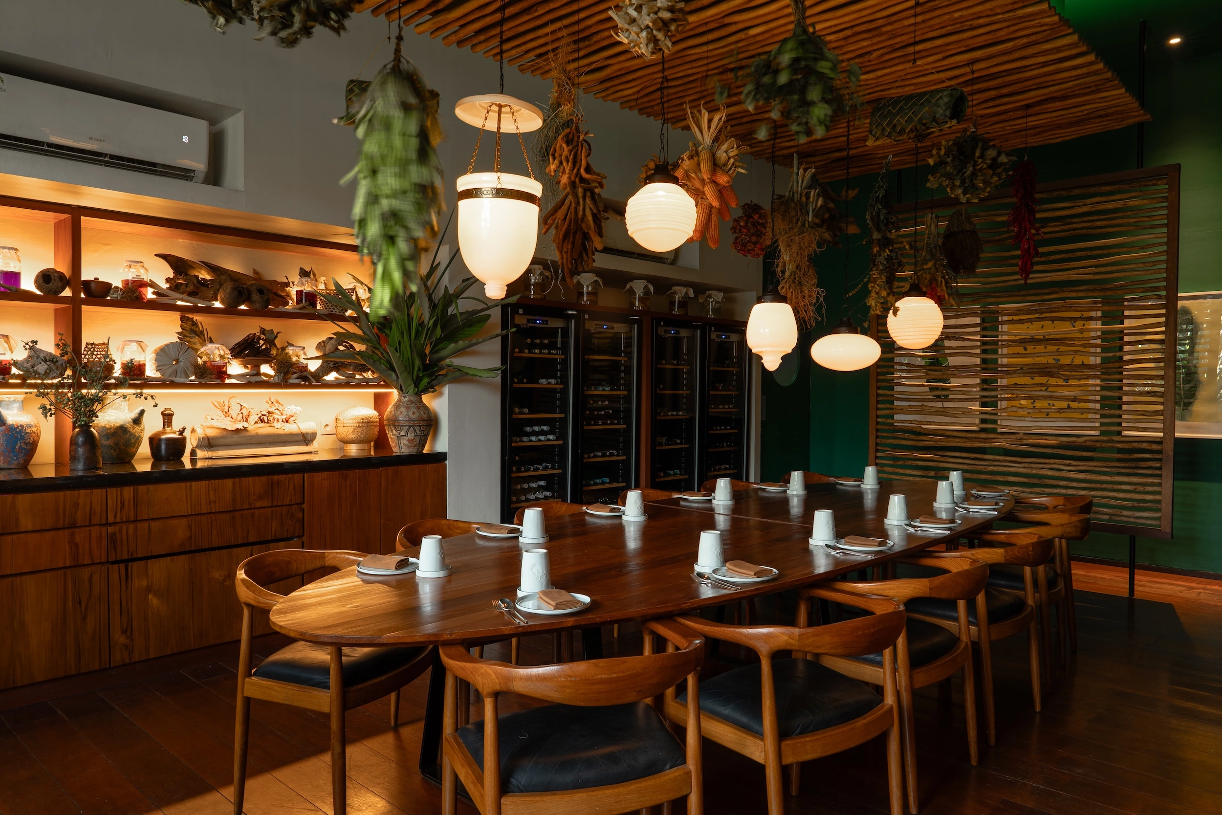 The private dining room — a long wooden table laid for a party under paper lanterns and hanging dried plants, with lit shelves of ceramics on one side and a wine wall behind