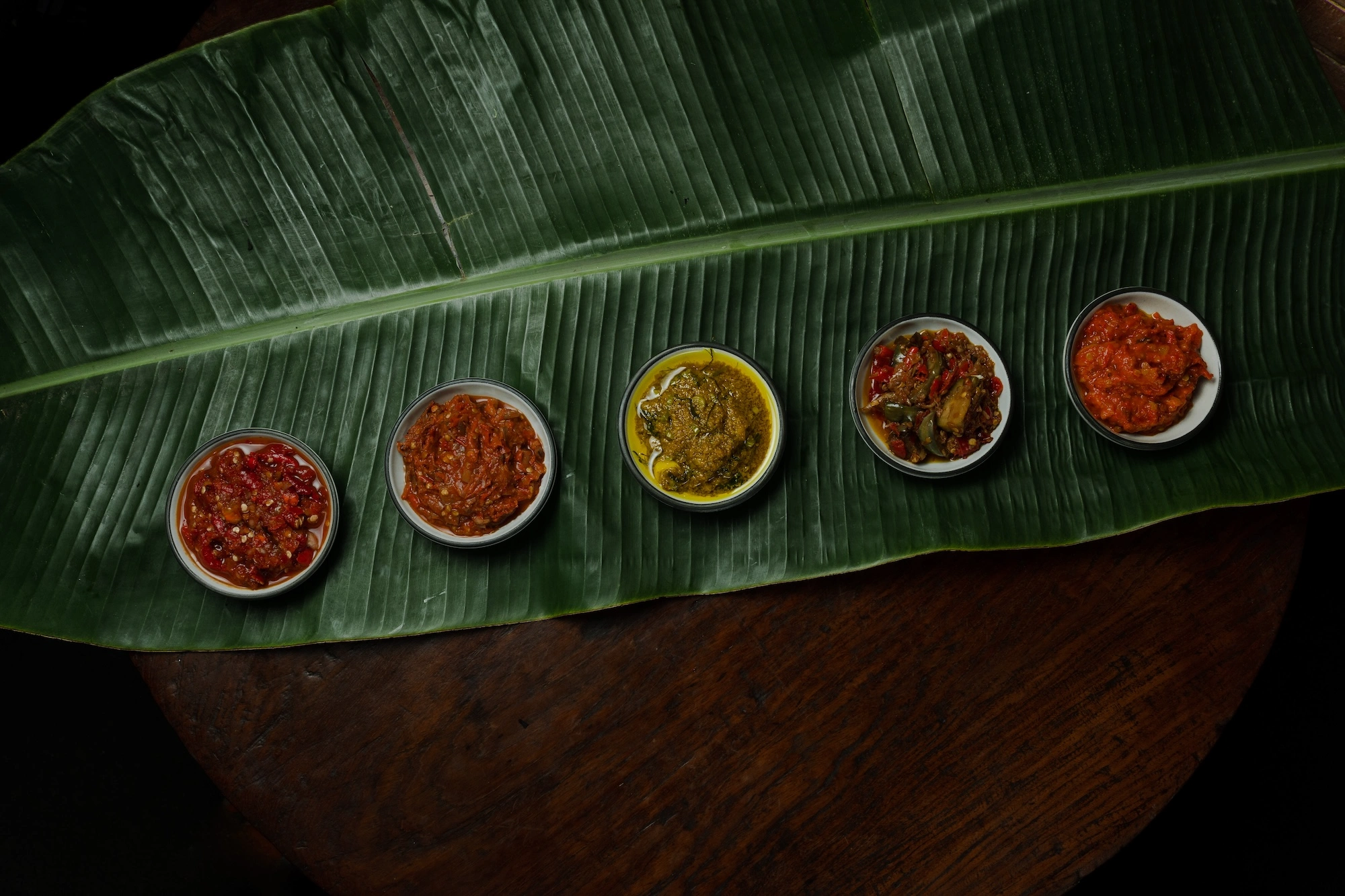 Five sambals in small bowls laid along a banana leaf