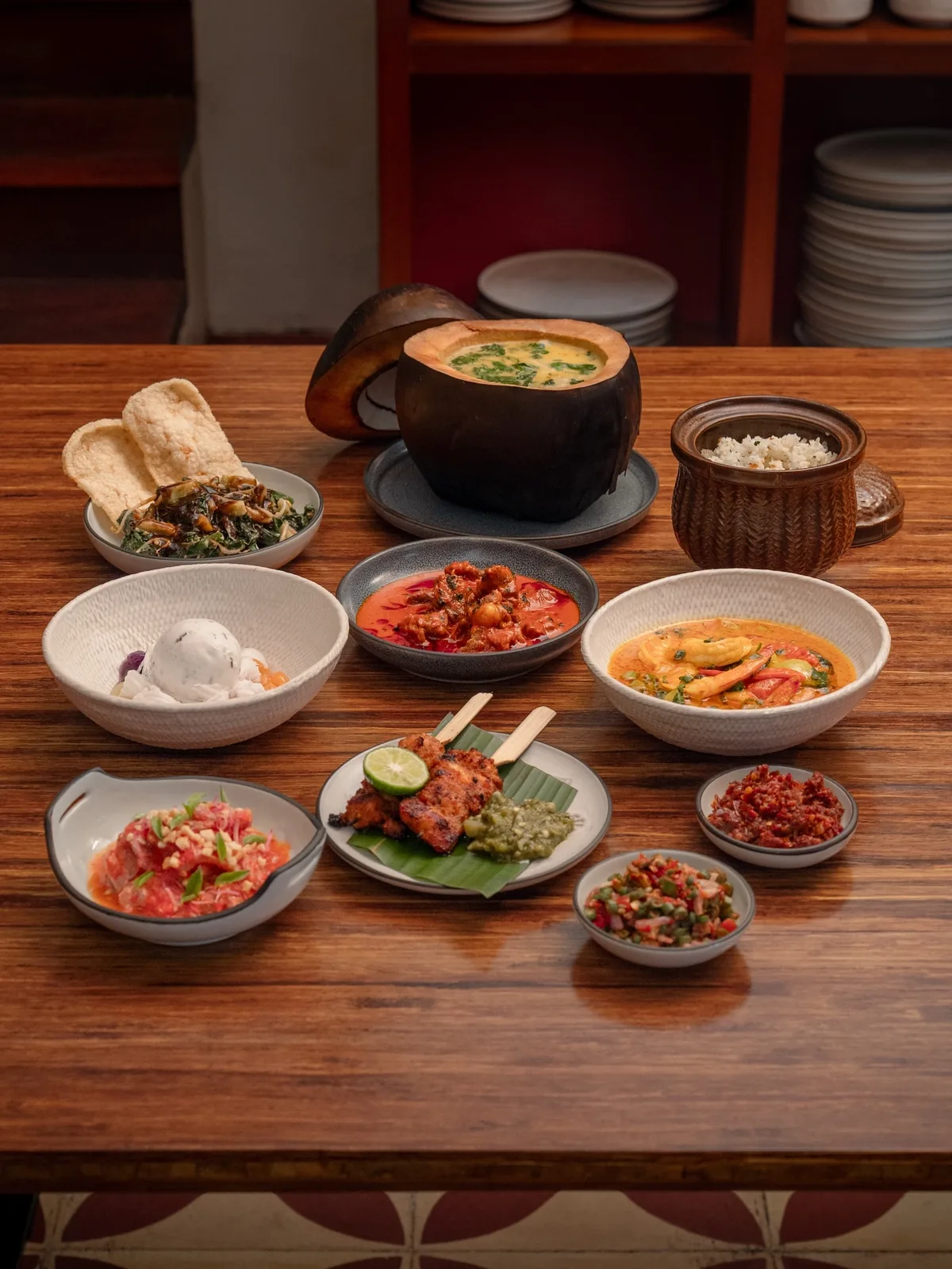 A set menu laid out along a wooden table — soup in a clay pot, rice, sate, sambals, curries and greens with crackers — with stacked plates on the shelves behind