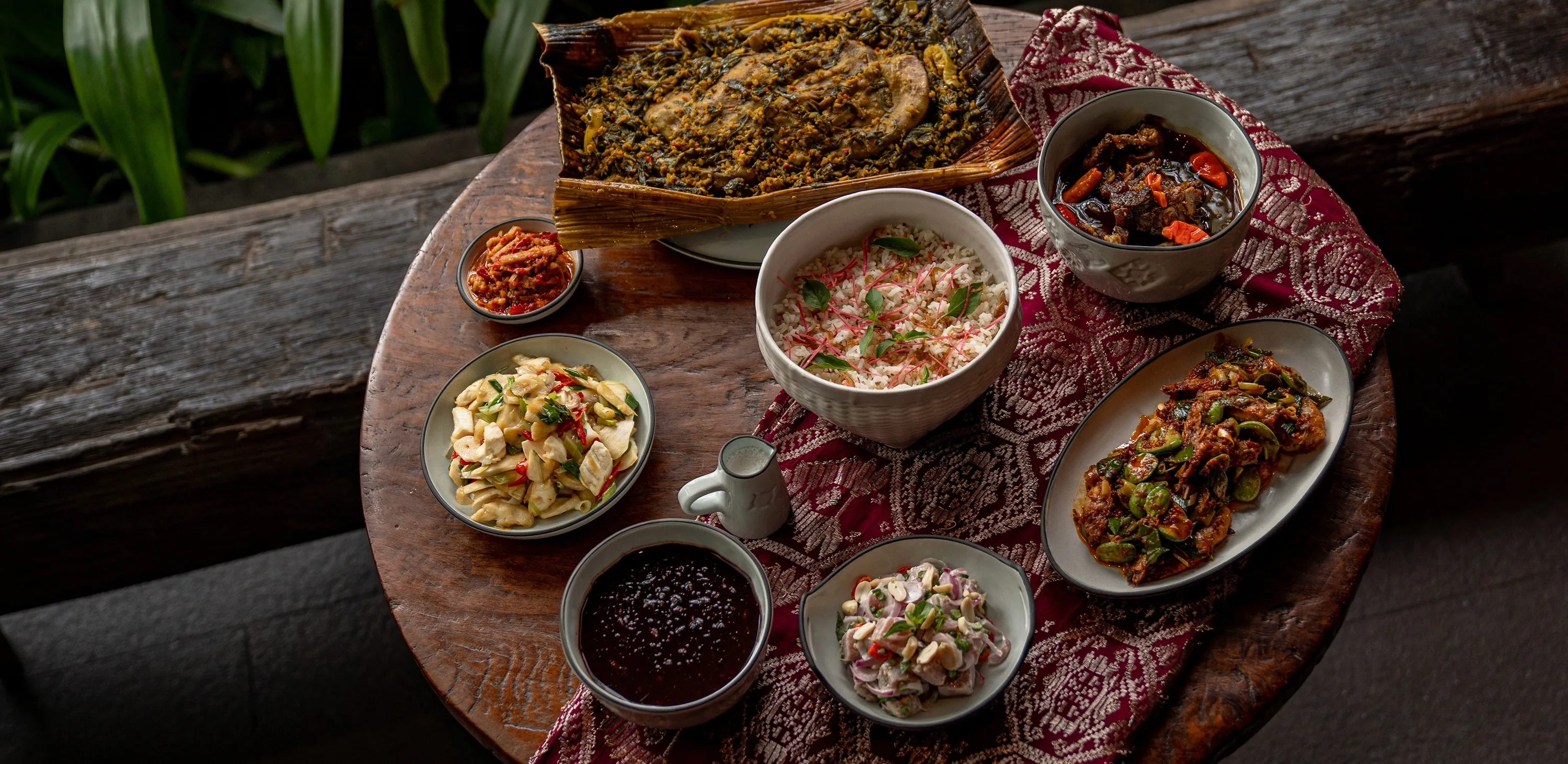The set menu on a round wooden table over a red songket cloth — a dish baked under greens in a palm sheath, herbed rice, a dark braise with chillies, petai in sambal, raw tuna with shallots and peanuts, a stir-fry with chilli and spring onion, and a purple sweet potato porridge with a jug of coconut milk
